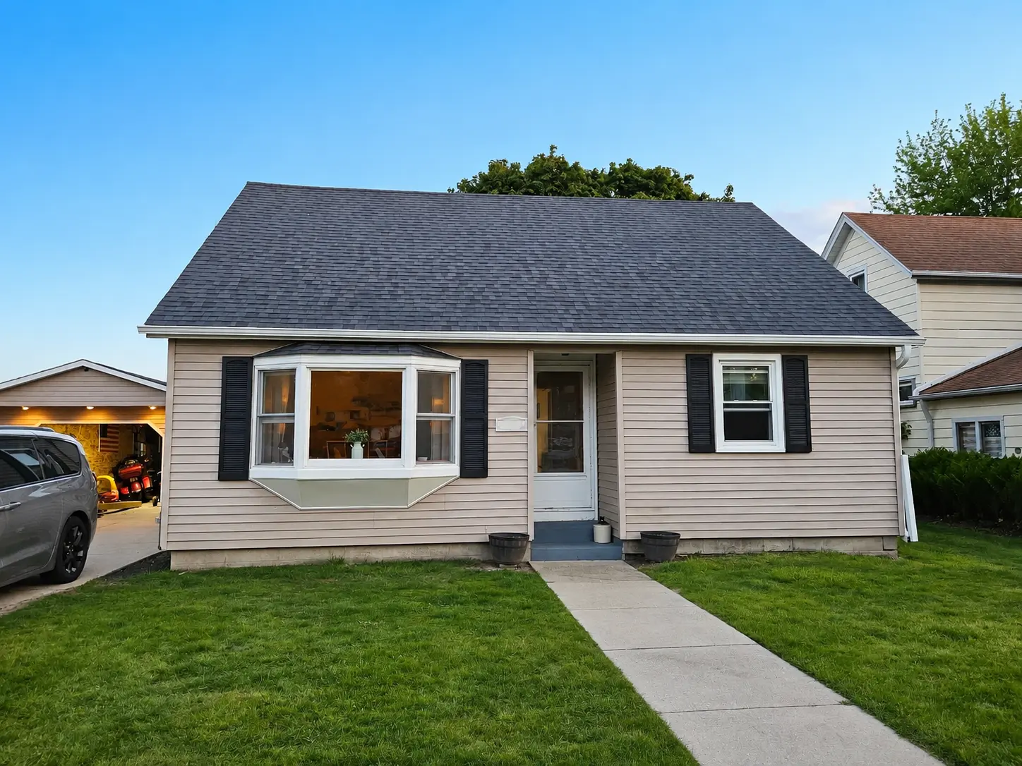 Front of a cream sided cape-style house with a new charcoal shingle roof and white fascia, photographed at dusk.